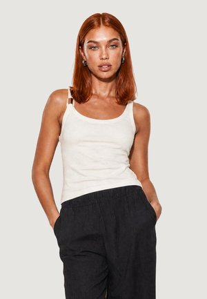 Donna con capelli rossi che indossa una canottiera bianca e pantaloni neri, in piedi con le mani in tasca davanti a uno sfondo semplice.