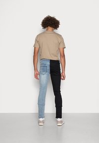 Men's two-tone jeans: one leg light blue denim, the other black denim. Features a casual beige t-shirt and sneakers. Back view.