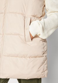 Krémový zateplený vest s hladkým povrchem, který má přední zip, dvě boční kapsy a ribbedmanžety rukávů vykukující zespodu.