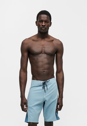Młody czarnoskóry mężczyzna stojący i noszący jasnoniebieskie spodenki kąpielowe ze sznurkiem na gładkim białym tle.