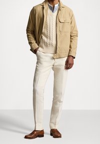 Muž v béžové bundě přes krémový svetr s pleteným vzorem, bílých kalhotách a hnědých kožených mokasínech, stojí se založen ýma rukama v kapsách.