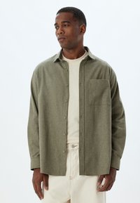 Olivgrüne Button-up-Bluse aus strukturiertem Stoff, mit einer Brusttasche und einer lockeren Passform, über einem cremefarbenen T-Shirt getragen.