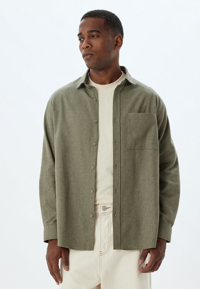 Olivgrüne Button-up-Bluse aus strukturiertem Stoff, mit einer Brusttasche und einer lockeren Passform, über einem cremefarbenen T-Shirt getragen.