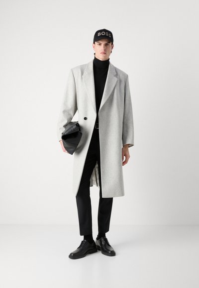 Manteau en laine gris avec un large col retour, pull à col roulé noir, pantalons noirs, baskets noires et casquette noire avec le logo "BOSS".
