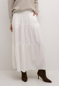 Bílá maxi sukně s volánkovým designem vyrobená z lehkého materiálu s elastickým pasem. Spárovaná s hnědými vysokými botami na podpatku.