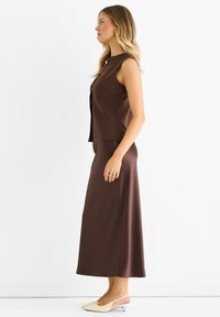 Brown sleeveless top with a high neckline, paired with a brown satin maxi skirt. Model wears beige slingback shoes with a low heel.