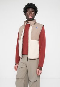 Beige and cream sleeveless vest with fleece front panels, zip closure, and logo. Paired with a red long-sleeve shirt and beige pants.
