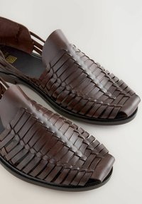 Sandalias de cuero marrón con parte superior trenzada, diseño de punta abierta y suela plana. Presentan una textura suave y bordes reforzados para mayor durabilidad.