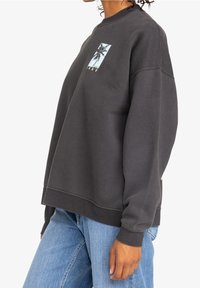 Felpa grigia con vestibilità rilassata, texture morbida e una grafica frontale che presenta palme. Maniche lunghe e polsini a coste. Abbinata a jeans azzurri chiari.