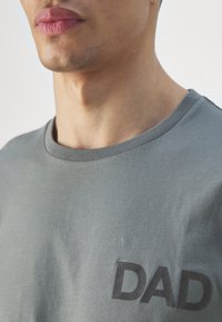 T-shirt en coton gris avec un col rond, arborant un imprimé noir "DAD" sur le côté gauche. Le tissu semble doux et légèrement texturé.