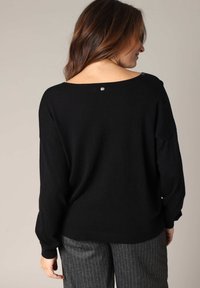 Pull en maille noir avec un décolleté large, des manches longues et un détail de bouton argenté au dos. Associé à un pantalon à rayures grises.