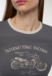 Zbliżenie na osobę w szarej koszulce z beżowymi wykończeniami oraz grafiką motocykla w stylu vintage z napisem "MIĘDZYNARODOWY TOR WYŚCIGOWY."