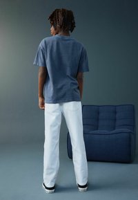 Blå t-shirt i bomuld med korte ærmer kombineret med hvide denimjeans og sorte sneakers, med synlig tekstureret sofa i baggrunden.