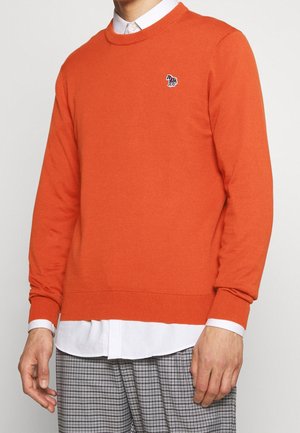 Mężczyzna w jaskrawo pomarańczowym swetrze crewneck na białej koszuli, w kratkowane spodnie, z małym haftowanym logo zwierzęcia na piersi.