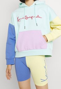 Persona que lleva una sudadera color bloque en tonos pastel con el texto "Kani Originals" y pantalones cortos a juego que presentan un contorno azul de una cara sobre tejido amarillo.