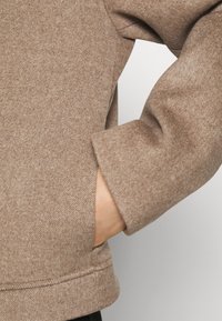 En persons hand i fickan på en beige ullrock med mjuk textur och långa ärmar.