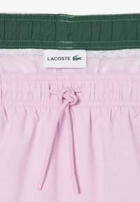 Maillot de bain rose avec ceinture élastique et cordon de serrage, doublure intérieure verte avec maille blanche, et étiquette logo Lacoste avec un crocodile.