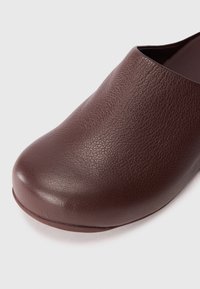 Mocassin en cuir marron avec un bout arrondi, une surface texturée et des détails lisses le long des bords ; design minimaliste.