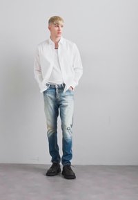 Una persona bionda in piedi contro un muro grigio, indossa una camicia bianca, una canottiera bianca, jeans blu usurati con una toppa e scarpe nere, con le mani in tasca.