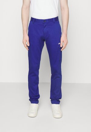 Pantalons cargo bleus à coupe ajustée, dotés de poches latérales et d'un bouton de fermeture. Confectionnés dans un matériau léger ; associés à des baskets blanches.