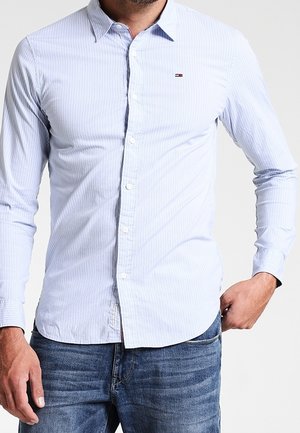Homme portant une chemise à rayures fines bleu clair boutonnée avec un petit logo sur la poitrine et un jean bleu, debout avec une main dans la poche.