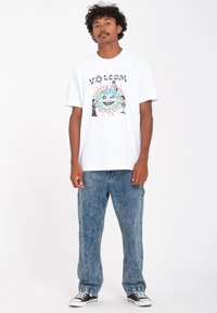 Witte katoenen t-shirt met een kleurrijke grafische print van verschillende figuren rond een wereldbol. Gecombineerd met verouderde blauwe denim jeans en zwarte sneakers.