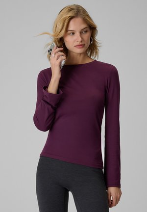 Blondynka w fioletowej bluzce z długim rękawem trzyma smartfon przy uchu, patrząc na bok na jednolitym jasnym tle.