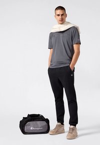 Szara koszulka z krótkim rękawem z haftowanym logo, czarne spodnie dresowe, beżowe sneakersy oraz czarna torba sportowa z szarymi akcentami.