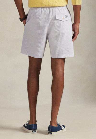 Άνδρας που φοράει ανοιχτό γκρι σορτς σερσάκερ με πίσω τσέπη, κίτρινο πουλόβερ και μπλε πάνινα παπούτσια, στέκεται μπροστά σε ουδέτερο φόντο.