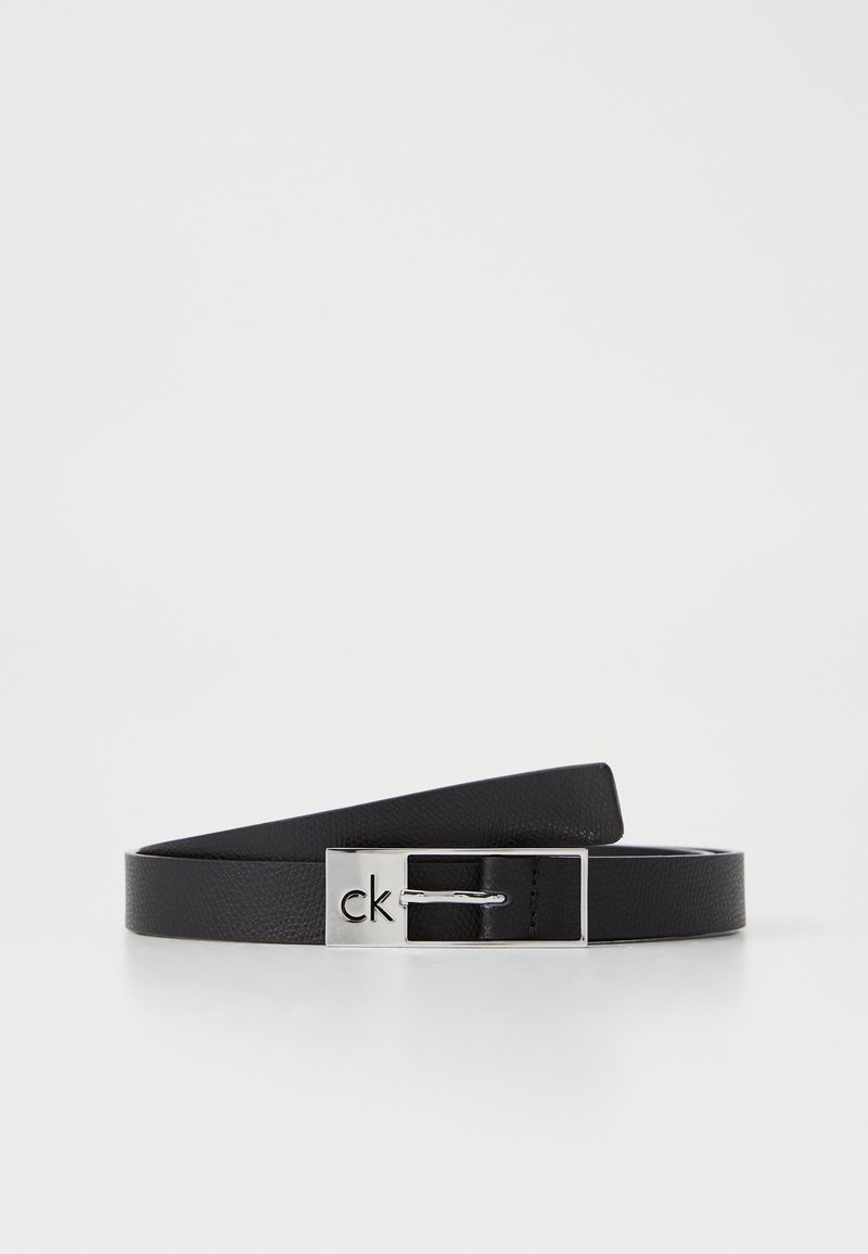 Ceinture en cuir noir avec une petite boucle rectangulaire en argent gravée du logo "ck", présentée enroulée sur une surface blanche.