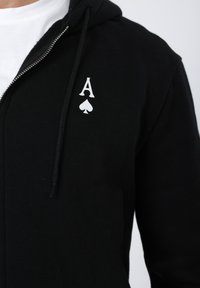 Μαύρο φούτερ με φερμουάρ από απαλό ύφασμα, με λευκό γραφικό που απεικονίζει ένα «A» και σύμβολο μπαστούνι στην αριστερή π λευρά του στήθους.