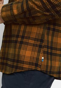 Orange und schwarzes kariertes Hemd mit abgerundetem Saum, geknöpftem Bündchen und einem kleinen blau-weißen Etikett an der Seitennaht. Weicher Stoff.