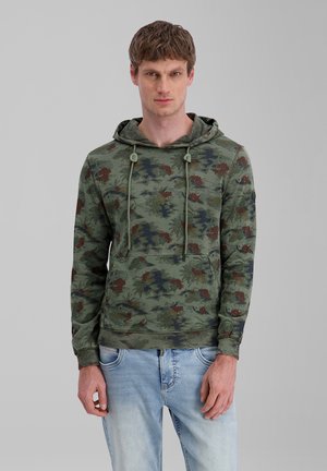 Junger Mann, der einen grünen Hoodie mit Camouflage-Muster und hellblaue Jeans trägt, stehend vor einem schlichten grauen Hintergrund.