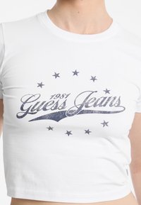 Balta apkirpta marškinėliai su tamsiai mėlynu blizgančiu logotipu "Guess Jeans" ir aplink juos esančiomis žvaigždėmis, pagaminti iš minkštos medvilnės trikotažo.