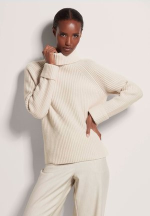 Ribbed turtleneck sweater in light beige, with long sleeves and relaxed fit. Made from textured knit fabric. Paired with light trousers.