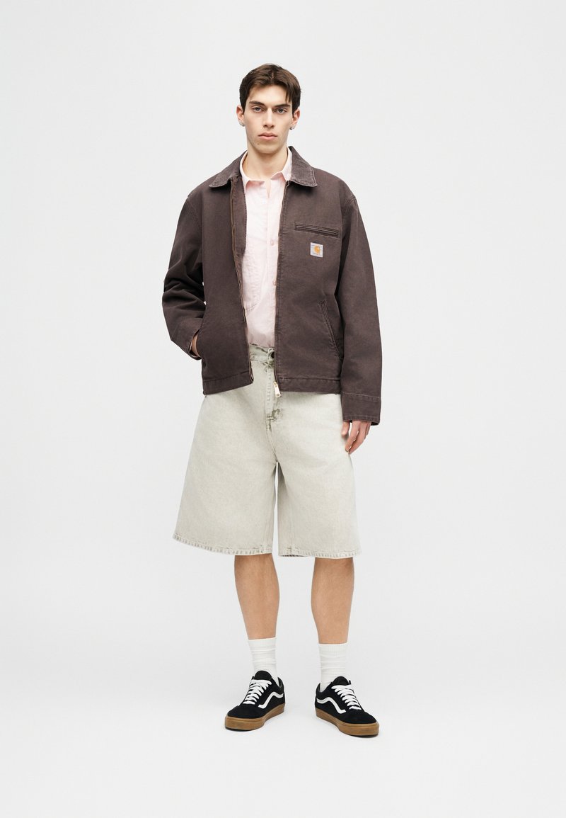 Joven con chaqueta marrón oscuro, camisa rosa claro, pantalones cortos beige hasta la rodilla, calcetines blancos y zapatillas negras, de pie frente a un fondo liso.