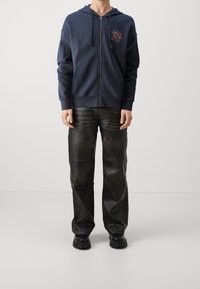 Felpa con zip navy, dotata di tasche frontali e ricamo di rose, abbinata a pantaloni in pelle nera e scarpe chunky nere con suola robusta.