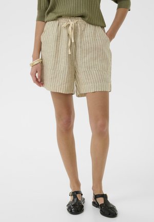 Frau trägt beige gestreifte Shorts mit Kordelzug, grünes geripptes Top, schwarze gewebte Sandalen und cremefarbene Armbänder am linken Handgelenk.
