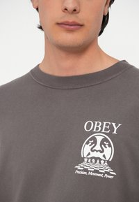Sivi dukserica s okruglim izrezom. Sadrži bijeli grafički otisak šahovske ploče i tekst "OBEY" iznad. Mekana tekstura.