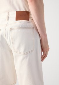 Des shorts en denim blanc présentent une taille haute, des coutures en cuivre et une étiquette en cuir brun au dos avec les détails de la marque.