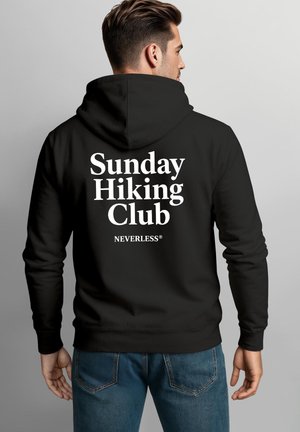 Man wearing black hoodie with hood down and white text "Sunday Hiking Club NEVERLESS" on the back, facing away against gray background.