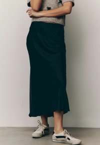 Gonna midi nera in satin con una silhouette fluida, abbinata a sneaker chiare con dettagli neri e una superficie testurizzata.
