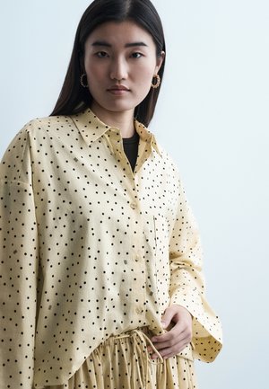 Kvinne med langt mørkt hår iført gullfargede hoops, gul skjorte med knapper og matchende skjørt med svarte prikker.