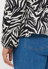 Blůza s černobílým vzorem zebry, s nabíranými manžetami, zkombinovaná s modrými džínami. Blůza má peplum střih a splývavou texturu.