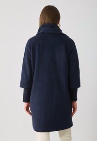 Marineblauer, samtig-texturierter Mantel mit hohem Kragen, weitem Ärmelschnitt und gerippten Manschetten. Schlichtes Design ohne sichtbare Verschlüsse oder Muster.