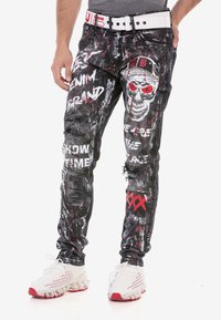 Schwarze, ausgewaschene Jeans mit roten und weißen Grafiken, darunter ein Schädelmuster und Text. Mit einem weißen Gürtel und sichtbaren Hardware-Details.