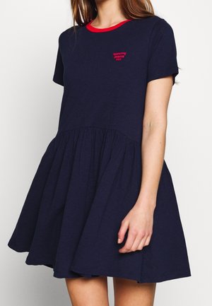 Woman wearing a navy blue short-sleeve dress with a gathered skirt and red trim at the neckline, featuring small red "tommy jeans" logo on chest.