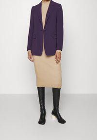 Fioletowa, dopasowana marynarka na beżowej, prążkowanej sukience, uzupełniona czarnymi kozakami za kolano z białymi bloczkowymi obcasami. Minimalistyczny design, strukturalne dopasowanie.