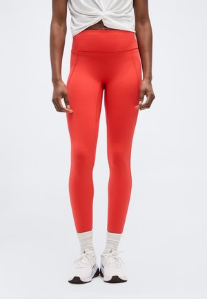 Kvinde iført røde leggings med høj talje, hvid vridd crop top, hvide sokker og hvide sneakers, stående med armene afslappet langs siden.