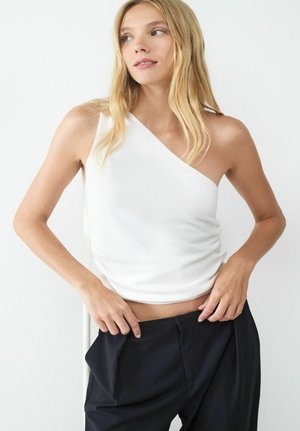 Vrouw met lang blond haar, gekleed in een witte top met één schouder en zwarte broek, die de tailleband aanpast en iets opzij kijkt.